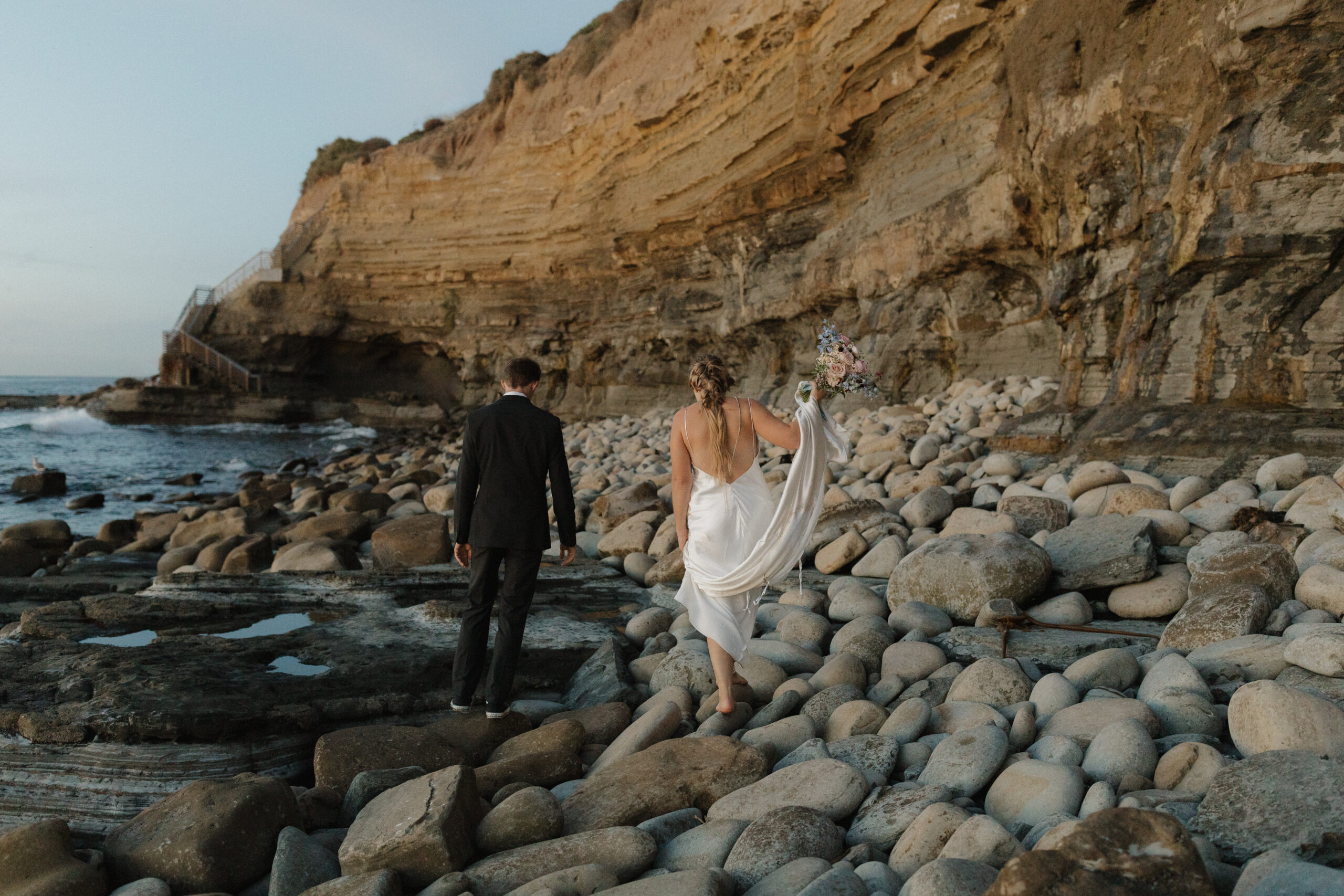 Bride and groom climbing across large beach rocks to get to the stairs